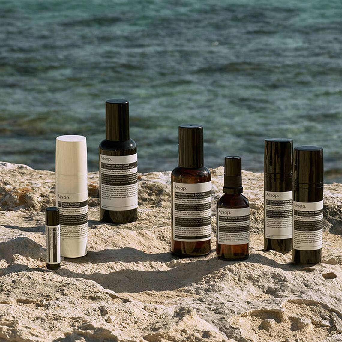 Eine sorgfaltige Auswahl an schutzenden Aesop Sonnenpflege-Begleitern, prasentiert auf einem Kustenfelsen am Meer.