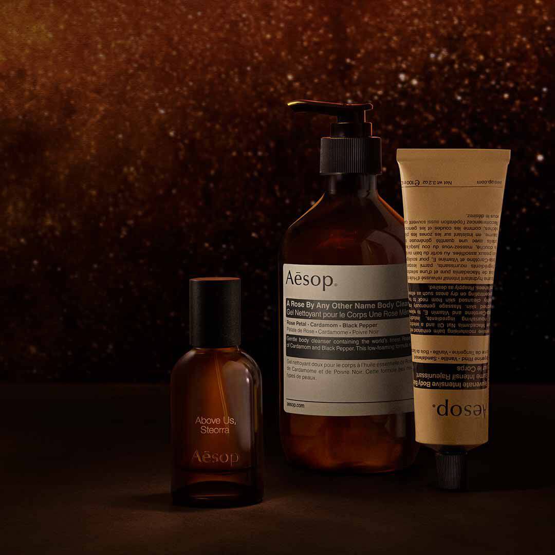 Aesop Above Us, Steorra Flakon mit Pumpspender und Tube vor einem warmen, braun gesprenkelten Hintergrund.