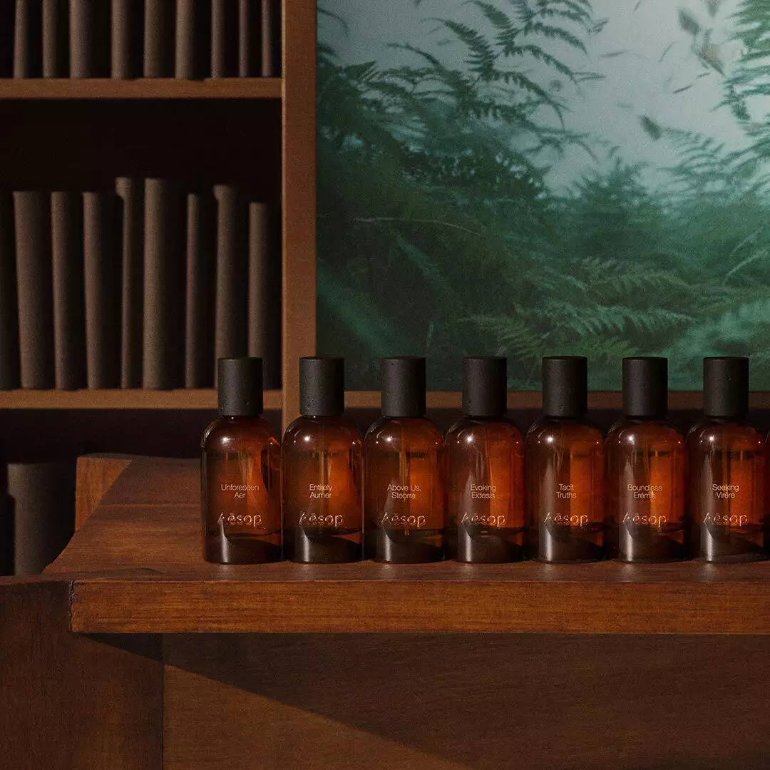 Reihe bernsteinfarbener Aesop-Duftflakons auf einem Holztisch neben Regalen und dem Bild eines grunen Farns.