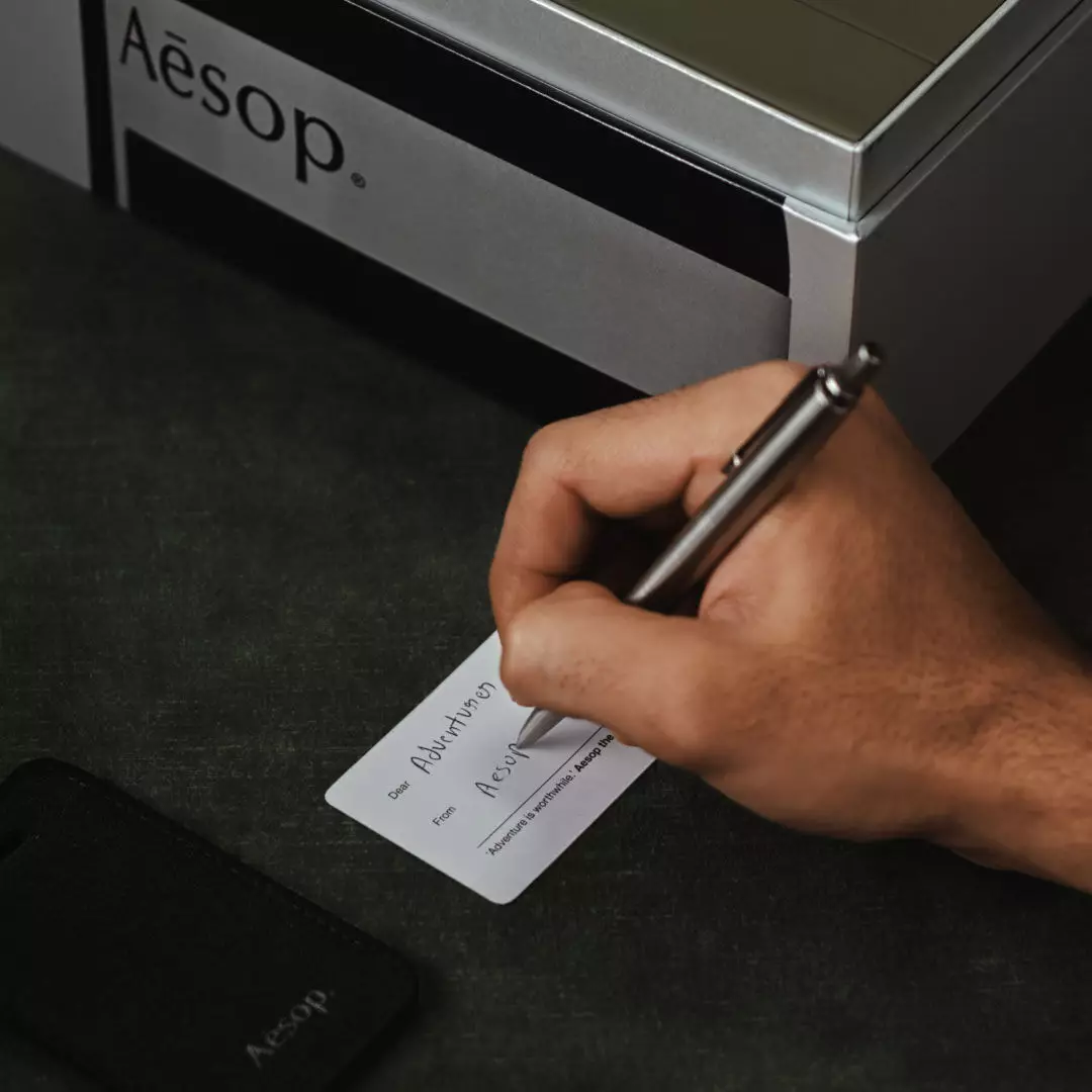 Eine Hand schreibt auf eine kleine weiße Karte neben einem schwarzen Aesop-Gepackanhanger und einem silbernen Aesop-Etui auf dunkelgrunem Stoff.