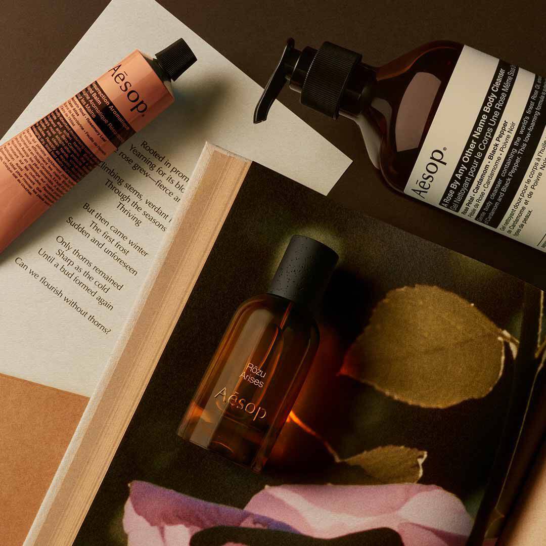 Aesop Rozu Arises Flakon mit Pumpspender und pfirsichfarbenem Schlauch, arrangiert auf Papieren mit rosafarbenen Blutenblattern und einem Blatt.