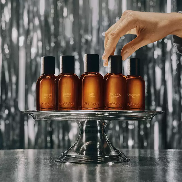 Eine Reihe von Aesop-Parfumflaschchen aus bernsteinfarbenem Glas auf einem Holzregal vor einer Bibliothek und einem grunen Kunstwerk.