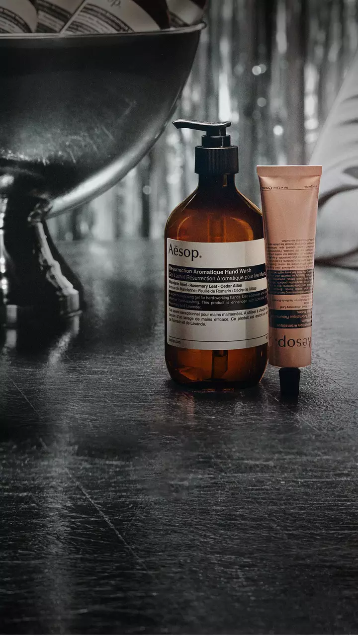 Eine bernsteinfarbene Aesop-Pumpflasche und eine rosafarbene Tube stehen neben einer großen Schale auf einem Sockel auf einem reflektierenden Tisch, dahinter silberne Girlanden.